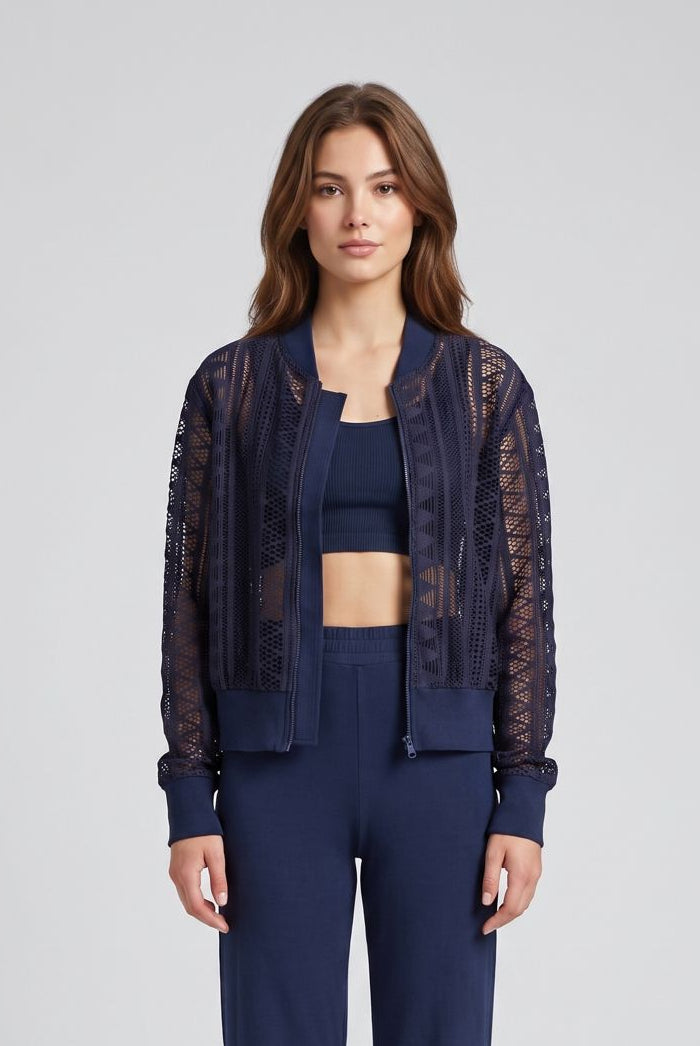 A woman stands barefoot against a light gray studio background wearing a navy blue two-piece lounge set with a cropped tank top and matching wide-leg pants, plus a sheer lace bomber jacket.